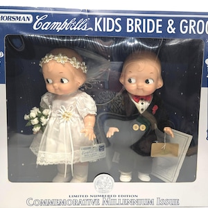 May include: A Horsman Campbell's Kids Bride & Groom doll set in a box. The bride wears a white dress, veil, and flower crown, holding a bouquet. The groom wears a black tuxedo with a red bowtie. The box is labeled Commemorative Millennium Issue.