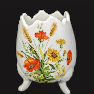 May include: White ceramic vase with three legs and a floral design. The vase features red poppies, yellow sunflowers, and wheat stalks.