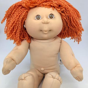 May include: A vintage doll with a soft, flesh-toned body and vibrant orange yarn hair. The doll has detailed facial features, including expressive eyes and a small earring. It is in a seated position.