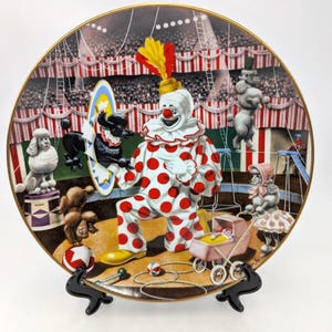 May include: Decorative plate featuring a circus scene with a clown in a red and white polka-dot suit. The plate has a gold rim and depicts poodles, a baby carriage, and a circus ring.