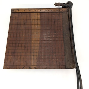 May include: A vintage paper cutter featuring a dark brown wooden base with a black grid pattern and a ruler along the top edge. The device includes a heavy-duty black metal cutting arm with a hinge mechanism and a long handle on the right side.