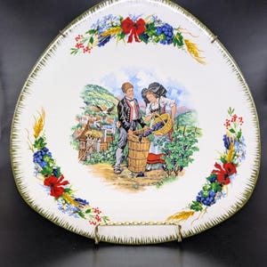May include: A white ceramic plate with a green rim and a painted scene of two people harvesting grapes in a vineyard. The plate is decorated with a floral border of red, blue, and white flowers.