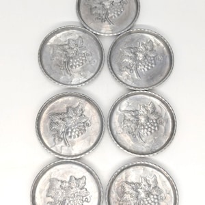 May include: A set of seven circular silver-toned metal coasters arranged in a pyramid formation. Each coaster features a raised embossed design of a cluster of grapes with leaves in the center and a decorative rope-style texture along the rim.