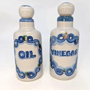 May include: Two off-white ceramic cruets with rounded stoppers. Each bottle features a hand-painted blue swirling border surrounding the text OIL on one and VINEGAR on the other. The designs include small green and peach accents within the blue.