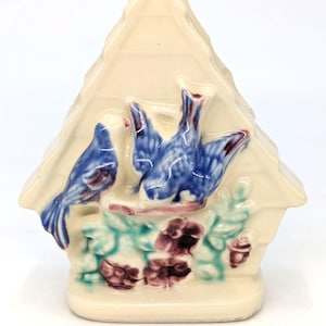 May include: A cream-colored ceramic napkin holder shaped like a house with a triangular roof. The front features a relief design of two blue birds perched on a branch above green leaves and dark purple flowers, all finished with a glossy glaze.