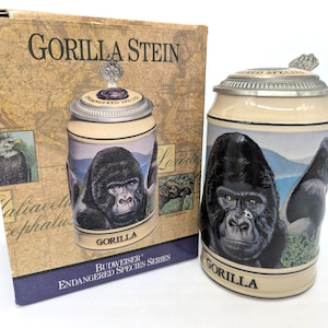 May include: A ceramic beer stein from the Budweiser Endangered Species Series featuring a detailed illustration of a gorilla, the word GORILLA printed at the base, and a metallic lid. It is displayed next to its original cardboard box packaging.