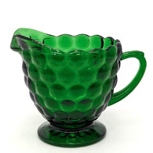 May include: A vintage green glass creamer with a hobnail pattern. The creamer has a handle and a spout, and sits on a round base. The glass is a deep emerald green color.
