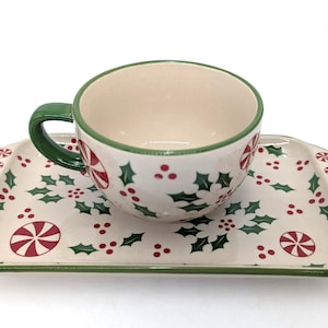 May include: A ceramic cup and rectangular tray set featuring a cream-colored base with a festive pattern of green holly leaves, red berries, and red and white peppermint candies. The cup has a green handle and a green rim, matching the tray's edge.