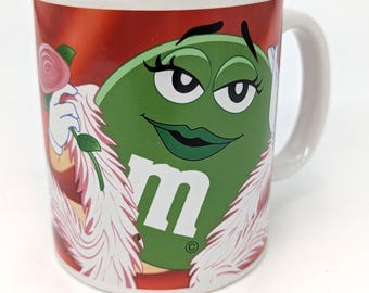 May include: White ceramic mug featuring a green M&M character with a glamorous design. The character has a green face, black eyelashes, and a red background. The mug has a white handle and a rose.