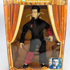 May include: An *NSYNC collectible marionette of Chris Kirkpatrick in a black jumpsuit with red accents, housed in a box featuring orange stage curtains, the text "Living Toyz," and a small portrait of the artist on the lower right corner.