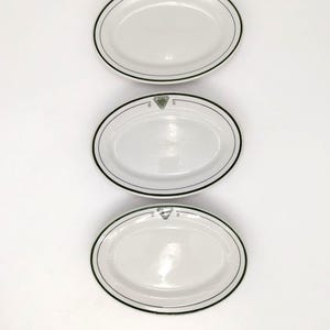 May include: Three white oval serving platters with a dark green border and a decorative emblem. The platters are arranged vertically against a white background. The plates are suitable for serving food.
