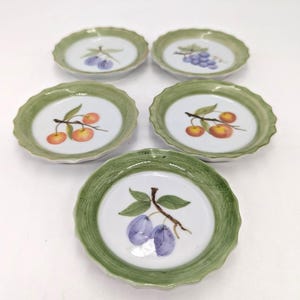 May include: A set of five small ceramic plates with scalloped edges and a green painted rim. Each plate features a central illustration of different fruits, including purple plums, blue grapes, and red cherries on branches against a white base.