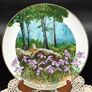 May include: A white ceramic plate with a gold rim features a painting of a forest scene with a fox sitting on a rock surrounded by purple flowers. The artist's signature is visible in the bottom right corner.