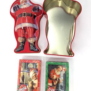 May include: A red Coca-Cola Santa Claus tin with an open lid, and two decks of Coca-Cola playing cards. The tin is shaped like Santa Claus. The playing cards are wrapped in plastic and feature Santa Claus.