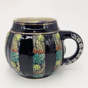 May include: A dark blue ceramic beer stein with a gold rim and handle. The stein features vertical panels with relief designs of hops and leaves in green, yellow, and orange. The handle has a white floral design.