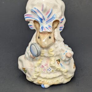 May include: A ceramic figurine of a mouse wearing a ruffled bonnet and a dress with blue polka dots and floral accents. The mouse holds a small circular hand mirror in one paw. The figurine features a glossy finish with painted details in white, blue, pink, and brown tones.