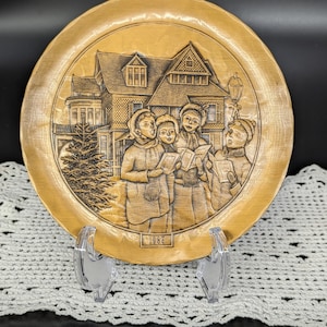 May include: A gold-toned metal plate with a raised design of a Victorian house and four carolers singing. The plate is dated 1986.