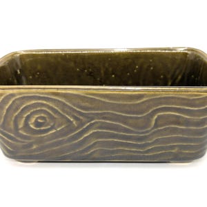 May include: A rectangular ceramic planter with a glossy olive green glaze. The exterior surface features a molded relief pattern that mimics the appearance of natural wood grain, complete with a prominent circular knot detail on the front side.