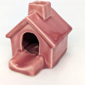 May include: A small, glossy, mauve-colored ceramic house figurine featuring a pitched roof, a central chimney, and a rounded front opening with a small, protruding entrance step. The surface has a smooth, reflective glaze finish throughout.