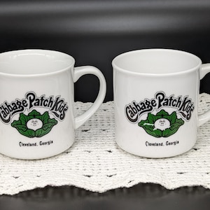 May include: Two white ceramic mugs with black and green Cabbage Patch Kids logos. The logos feature a green cabbage with a white face and the text "Cabbage Patch Kids" in a black arch above the cabbage. The text "Cleveland, Georgia" is printed below the cabbage.
