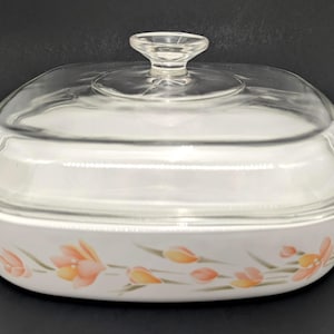 May include: A white rectangular casserole dish with integrated side handles and a clear glass lid featuring a knob handle. The exterior of the dish is decorated with a horizontal pattern of soft orange and yellow tulip-like flowers with green stems.