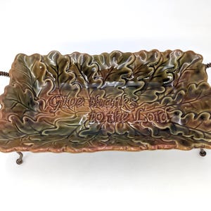May include: A rectangular ceramic serving dish with a glossy finish in shades of brown and green, shaped like a cluster of oak leaves. The center features the embossed text "Give thanks to the Lord." It has two curved, textured metal handles.