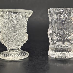 May include: Two clear glass vases. The vase on the left has a flared top, a bulbous midsection, and a pedestal base. The vase on the right is cylindrical with a textured, stacked design. Both are transparent.