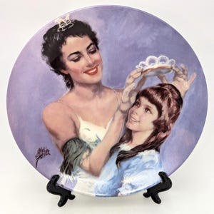 May include: A decorative plate featuring a painting of a woman placing a tiara on a young girl's head. The plate has a purple background and is supported by a black stand. The artwork is signed by Shell Fisher.