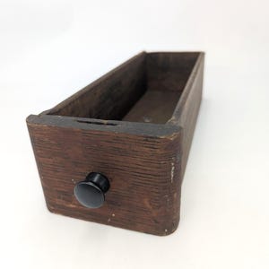 May include: A rectangular wooden drawer with a dark brown, weathered finish and a visible wood grain texture. The drawer features a single, small, round, black knob centered on the front panel. The interior is hollow and open at the top.