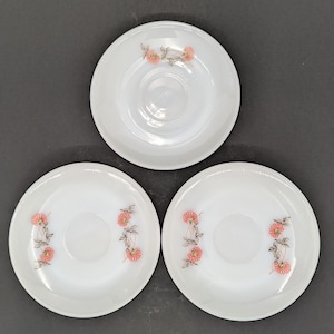 May include: Three white saucers with a floral design. Each saucer features a central depression and a delicate pattern of pink flowers and green leaves. The saucers are arranged on a dark surface.