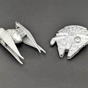 May include: Two silver-colored metal miniature starship models. One model is a TIE interceptor with angular wings and a central cockpit. The other model is a circular, flat-topped spacecraft with intricate surface detailing and a side-mounted cockpit.
