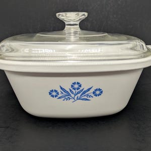 May include: White casserole dish with a clear glass lid. The dish features a blue floral design on the front. The lid has a clear glass handle. The dish is designed for cooking and serving.