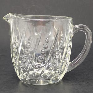 May include: Clear glass creamer with a handle and a spout. The creamer has a textured design of diagonal lines and small circles. The creamer is likely vintage and suitable for serving cream or other liquids.