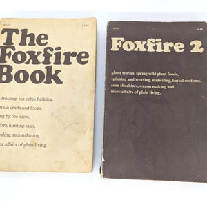 May include: Two vintage books, "The Foxfire Book" and "Foxfire 2", are displayed on a white surface. The first book has a cream-colored cover with brown text, while the second has a dark brown cover. Both books feature detailed text about plain living.