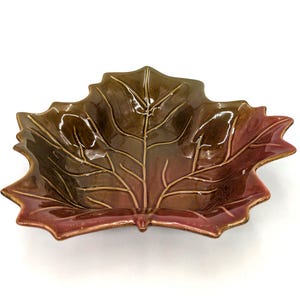 May include: A ceramic bowl shaped like a maple leaf with a glossy finish. The bowl features a gradient color scheme transitioning from deep brown at the center to a reddish-maroon hue at the edges, with raised, light-colored vein details.