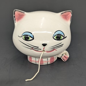 May include: A ceramic cat-shaped yarn bowl with a glossy off-white finish and a fine crackle glaze. The cat features pink-lined ears, bright green eyes with blue eyeshadow, black whiskers, and a pink mouth. A pink and white plaid collar surrounds the base.