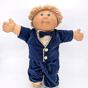 May include: A doll with tan skin, green eyes, and curly light brown yarn hair. The doll wears a dark blue velvet suit jacket with two white buttons, a matching bowtie, and dark blue velvet trousers over a cream-colored lace-trimmed shirt.