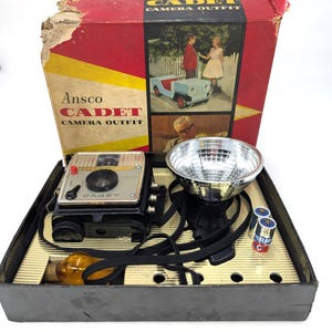 May include: An Ansco Cadet camera outfit in a red and black cardboard box. The set includes a black box camera, a silver metallic flash reflector, a yellow light bulb, and two Eveready batteries, all arranged in a gold-toned plastic insert.