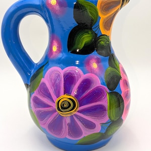 May include: A hand-painted blue ceramic pitcher with a large purple flower and green leaves. The pitcher has a curved handle and is decorated with additional pink and orange floral accents. The design is vibrant and colorful, perfect for serving beverages.