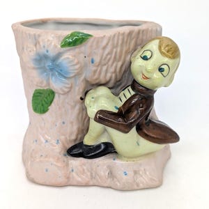May include: A vintage ceramic planter in the shape of a tree trunk, painted pink with blue accents and a blue flower. A seated figurine in a brown jacket and black shoes is leaning against the planter. The planter is a unique decorative item.