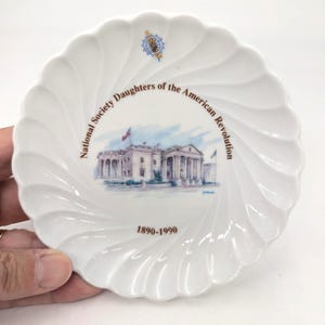 May include: A white porcelain scalloped plate featuring a central illustration of a building with columns, the text "National Society Daughters of the American Revolution," a small blue and gold emblem at the top, and the dates "1890-1990" at the bottom.