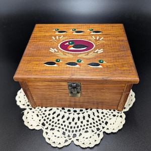 May include: A wooden box with a hinged lid and a metal latch. The top of the box is decorated with a painted scene of ducks in a pond. The ducks are painted in shades of green, black, orange, and red.