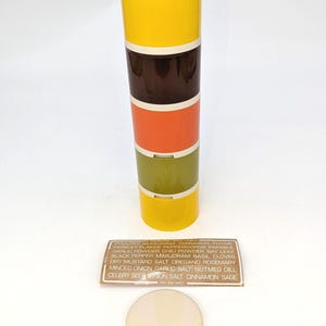 May include: A multi-colored spice container with a cream-colored lid. The container is stacked with yellow, brown, orange, and green sections. A label lists various spices. The lid is separate, and the container is on a white background.