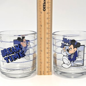 May include: Two clear glass mugs with handles, each featuring a cartoon image of Mickey Mouse in a blue suit. One mug has the words "BREAK TIME" in blue. The mugs have blue horizontal lines and are approximately 3 inches tall.