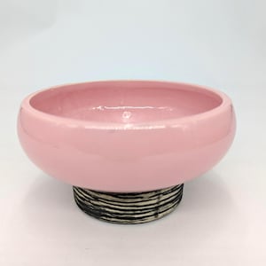 May include: A round ceramic bowl with a smooth, glossy light pink interior and exterior finish. The bowl is supported by a circular base featuring a textured, horizontal black and white striped pattern that contrasts with the solid pink upper section.