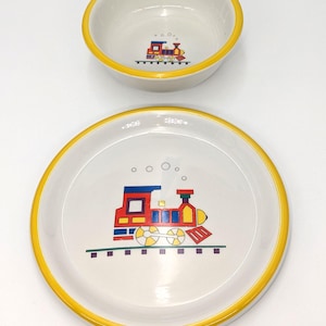May include: A white ceramic bowl and plate set with a colorful train design. The train features red, yellow, blue, and green accents. Both pieces have a bright yellow rim. The plate measures approximately 8 inches in diameter.