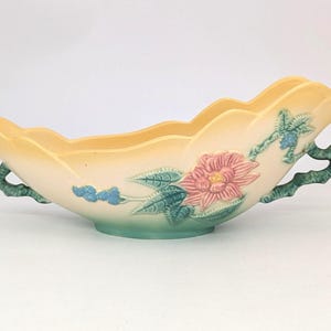 May include: A vintage, oblong ceramic planter with a scalloped rim. The planter is cream-colored with a yellow interior and features a pink and green floral design. It has green, branch-like handles and measures approximately 12 inches long.