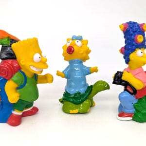 May include: Three Simpsons figurines: Bart with a backpack, Lisa riding a turtle, and Marge holding a camera and a red book. The figures are yellow with colorful clothing and accessories, set against a white background.