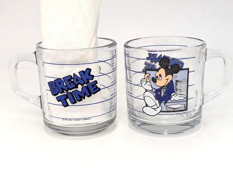 May include: Two clear glass mugs with handles. One mug has the words "BREAK TIME" in blue and white. The other mug features a cartoon image of Mickey Mouse in a blue suit. Both mugs have blue horizontal lines.