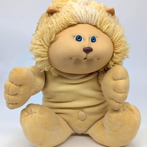 May include: A vintage plush doll in the shape of a lion cub. The doll has a tan face with blue eyes and a small nose. It has a yellow yarn mane and is wearing a tan onesie. The doll is sitting on a white surface.